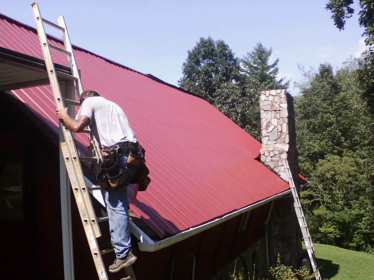 Roofer on a ladder beside a red metal roof and stone chimney — Soffit & Fascia Repair by Roofing Brothers in Davidsville